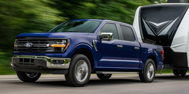 Blue Ford F-150 pickup truck towing a large travel trailer on a road with blurred green background.