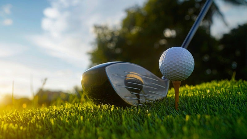 Golf ball on a tee with a golf driver ready to swing on green grass, bathed in golden sunlight.