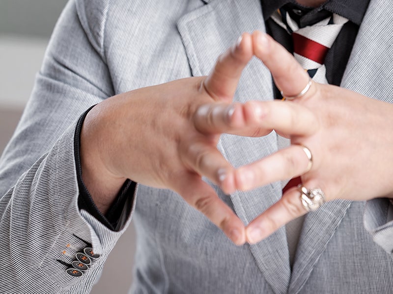 Hands interlocked in a triangular shape, wearing rings, a pinstriped blazer, and a striped tie.